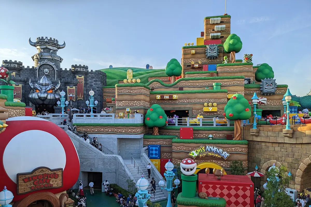 Part of Super Nintendo World, with a dark stone castle at the left, a green stepped hillside with trees and a red mushroom building in front